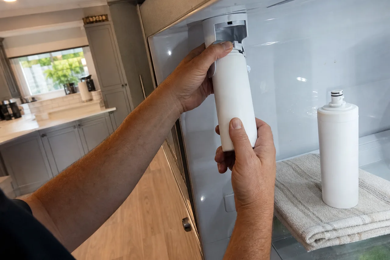 Two hands drawing a white filter cartridge down out of its housing at the top of a refrigerator compartment, a fresh cartridge waiting on a folded towel beside them.